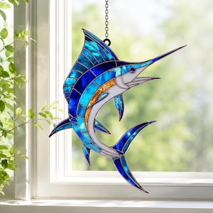 May include: A stained glass suncatcher shaped like a marlin fish, featuring vibrant shades of blue, turquoise, and amber glass pieces held together by dark metal soldering. The fish is depicted in a leaping pose and hangs from a metal chain.