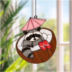 May include: A stained glass suncatcher featuring a gray and black raccoon sitting inside a brown coconut shell. The raccoon is positioned under a pink cocktail umbrella and has a pair of red flip-flops resting beside it. The piece hangs by a chain.