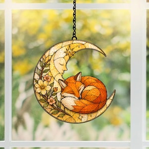 May include: A stained glass suncatcher featuring a sleeping fox curled up in a crescent moon. The moon is yellow with a face, stars, and floral accents. The fox is orange and white, with a black chain for hanging.