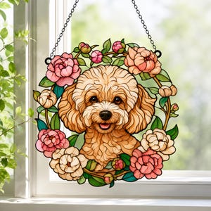 May include: A circular stained glass suncatcher featuring a light brown curly-haired dog surrounded by a wreath of pink, peach, and cream-colored peony flowers with green leaves. The piece hangs from a black metal chain against a window.