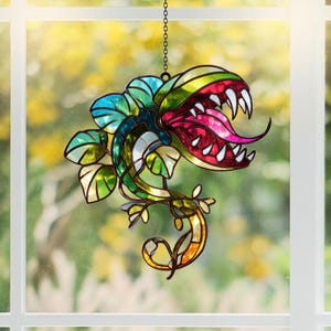 May include: A stained glass suncatcher shaped like a carnivorous plant with a wide-open mouth, sharp white teeth, and a long pink tongue. The plant features vibrant green, blue, and yellow glass panels with dark metal soldering, hanging by a chain.