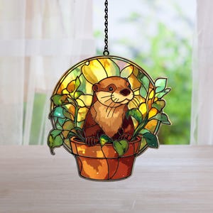 May include: A stained glass-style suncatcher depicting a brown otter in a terracotta-coloured pot surrounded by green leaves and yellow flowers. The otter is smiling. The suncatcher hangs from a black chain.