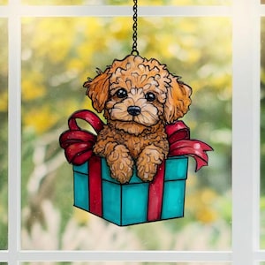 May include: A decorative window hanging featuring a brown poodle puppy inside a teal gift box with a red bow. The puppy has big black eyes and is suspended by a chain. The background is a blurred view of a green and yellow outdoor scene.
