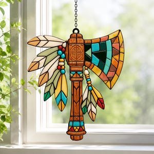 May include: A stained glass suncatcher shaped like a tomahawk with a brown wood-grain handle, a geometric blade in shades of teal, orange, and black, and decorative hanging feathers in white, yellow, teal, and red, suspended by a metal chain.