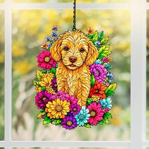 May include: A decorative hanging ornament featuring a golden-colored dog surrounded by colorful flowers. The flowers include shades of pink, purple, yellow, red, and blue. The ornament hangs from a black chain and is set against a blurred background.