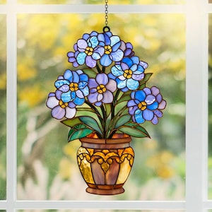 May include: A stained glass suncatcher featuring a bouquet of blue and purple flowers with yellow centers in a brown and amber pot. The piece is suspended by a metal chain and displays intricate glass segments with dark leaded outlines.