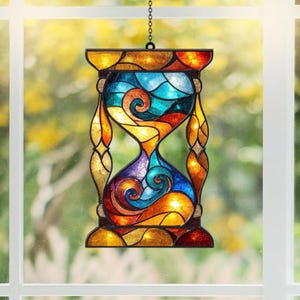 May include: A stained glass suncatcher shaped like an hourglass, featuring a vibrant mosaic of blue, orange, yellow, and purple glass pieces held together by dark metal framing, suspended by a thin chain against a bright window background.