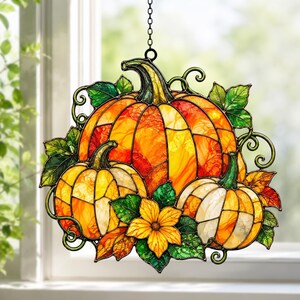 Puede incluir: Un atrapasoles de vidrieras con tres calabazas en tonos naranja, amarillo y crema, acentuado con hojas verdes, zarcillos y una flor amarilla central. La pieza está suspendida por una cadena de metal y presenta líneas de plomo oscuras.