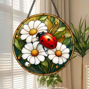 May include: A circular stained glass suncatcher featuring a vibrant red ladybug with black spots resting on white daisy flowers with yellow centers. The design includes green leaves and a dark metal frame, suspended by a black chain for display.