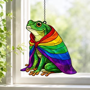 Könnte beinhalten: Ein Buntglas-Suncatcher, der einen grünen Frosch mit einem leuchtenden, regenbogenfarbenen Umhang zeigt. Der Frosch hat große, dunkle Augen und sitzt in einer aufrechten Position. Das Objekt hängt an einer dünnen Metallkette.