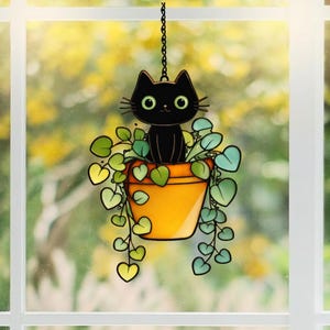 May include: A black cat with green eyes sits in an orange flower pot with green and yellow trailing leaves. The cat and pot are hanging from a chain, creating a whimsical, decorative piece.
