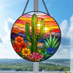 May include: A circular stained glass suncatcher featuring a desert landscape with a tall green saguaro cactus, agave, and colorful flowers in shades of orange, yellow, and pink against a vibrant sunset sky with purple mountains and a yellow sun.