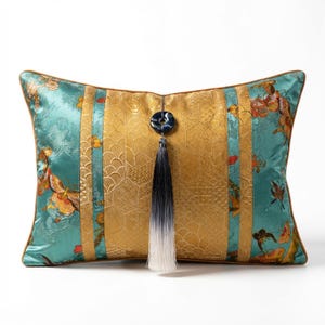 May include: Decorative rectangular pillow with a teal and gold floral pattern. The pillow features a gold center panel with a geometric design, teal side panels, and a dark blue tassel with a decorative button.