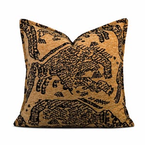 Puede incluir: Un cojín cuadrado con una base de tela texturizada de color bronce dorado y un patrón abstracto negro prominente. El diseño consiste en formas y líneas orgánicas estilizadas que se repiten, creando un visual denso y de alto contraste.