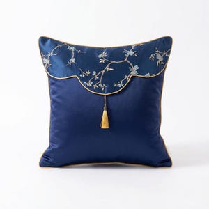 May include: A navy blue decorative pillow with a scalloped top section featuring embroidered floral branches in light blue. The pillow has a gold trim and a gold tassel. The pillow is square and made of a smooth fabric.