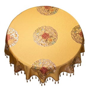 Könnte beinhalten: Ein runder, goldgelber Tischläufer mit verzierten, kreisförmigen Mustern in Rot, Gold und Creme. Der Tischläufer hat einen gewellten Rand mit kleinen Quasten. Das Design zeigt traditionelle asiatische Motive.