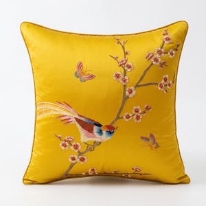 Può includere: Un cuscino decorativo quadrato giallo con un design ricamato. Il design presenta un uccello colorato appollaiato su un ramo fiorito, con farfalle. Il cuscino ha un bordo dorato.