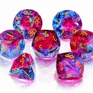 May include: A set of seven polyhedral dice featuring a vibrant gradient of magenta, purple, and blue hues with embedded iridescent flakes. Each die has a faceted, gem-like surface and is inscribed with clear, gold-colored numbers for readability.