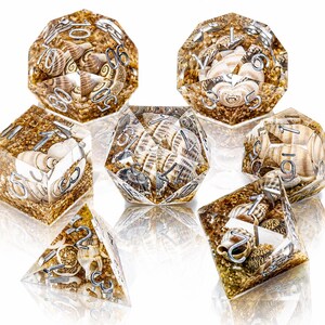 May include: A set of seven polyhedral dice made of clear resin, each containing small seashells and golden sand particles. The dice feature silver-colored numbers etched into the surfaces, including a d20, d12, d10, d8, d6, d4, and a percentile die.