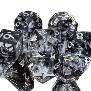 May include: A set of clear and black marbled polyhedral dice with silver numbering. The dice are in various shapes, including a d4, d6, d8, d10, d12, d20, and a percentile die. The dice are arranged on a white background.