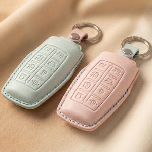 Op de afbeelding: Twee leren sleutelhangers in pastelkleuren, &eacute;&eacute;n lichtblauw en &eacute;&eacute;n roze, met zilveren sleutelringen. Elke sleutelhanger heeft reli&euml;fknopen en een bijpassende leren riem. De sleutelhangers zijn gestikt met een contrasterende draad.