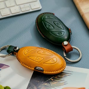 Op de afbeelding: Twee leren sleuteletuis in groen en geel-oranje, met zilveren sleutelringen. De groene hoes heeft een textuurpatroon en een bruine riem. De geel-oranje hoes heeft in reliëf aangebrachte sleutelsymbolen.