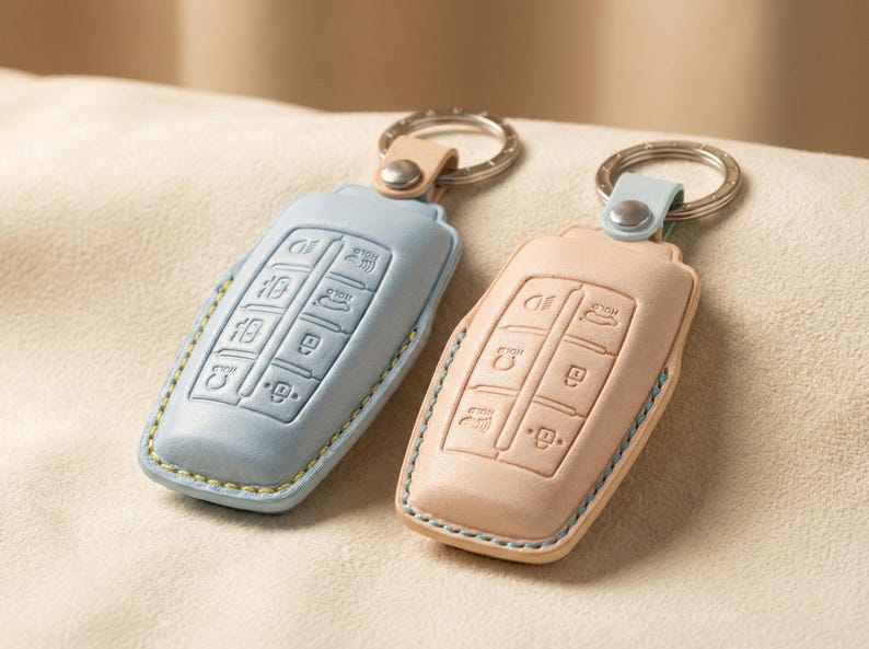 Op de afbeelding: Twee leren sleutelhangers in pastelkleuren. E&eacute;n is lichtblauw, de andere perzik. Beide hebben een rechthoekige vorm met afgeronde randen en reli&euml;f sleutelsymbolen. Elke sleutelhanger heeft een metalen ring en een bijpassende leren lus.