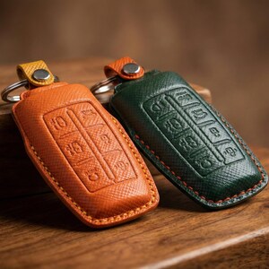 Op de afbeelding: Twee leren sleuteletuis, &eacute;&eacute;n oranje en &eacute;&eacute;n donkergroen, met zilveren sleutelringen. De oranje hoes heeft een gele accentkleur en de groene hoes heeft een oranje accentkleur. Beide hoezen hebben reli&euml;fknopontwerpen.