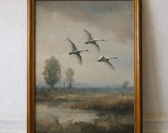 May include: An antique framed painting depicts three white swans in flight against a cloudy sky. The landscape features a marshy area with trees and a body of water, all rendered in muted tones.