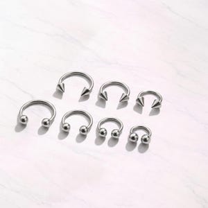 May include: A selection of silver horseshoe-shaped body jewellery. Some pieces have rounded ends, while others have pointed cone-shaped ends. The jewellery is arranged on a white marble-effect surface.