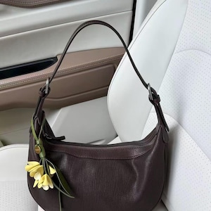 May include: A dark brown leather shoulder bag with a curved shape. The bag has a single strap and a small floral charm with yellow flowers and green leaves. The bag is set against a white and tan background.