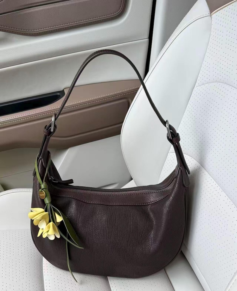 May include: A dark brown leather shoulder bag with a curved shape. The bag has a single strap and a small floral charm with yellow flowers and green leaves. The bag is set against a white and tan background.