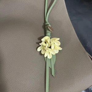 May include: A light green leather bag charm with a cluster of pale yellow flowers and a green leather strap. The charm is attached to a taupe leather bag.
