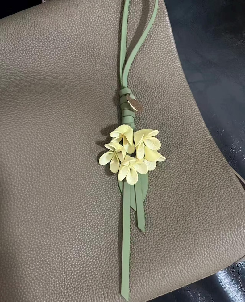 May include: A light green leather bag charm with a cluster of pale yellow flowers and a green leather strap. The charm is attached to a taupe leather bag.