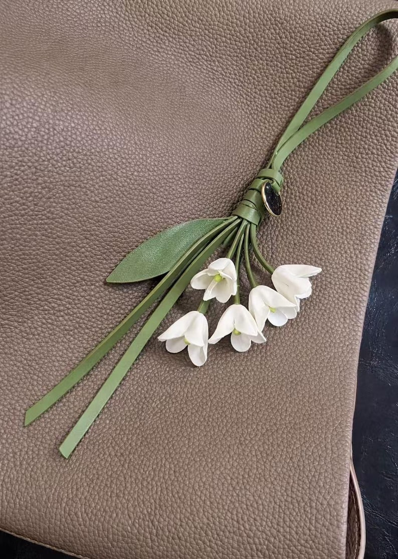 May include: A leather floral accessory featuring white bell-shaped flowers, green leaves, and a green strap. The accessory is attached to a textured, brown leather surface. The flowers have a delicate appearance.