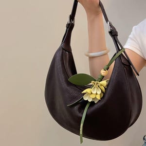 May include: A dark brown crescent-shaped handbag with a textured surface. The bag has a long shoulder strap and a decorative green and yellow floral accent. The bag is held by a person.