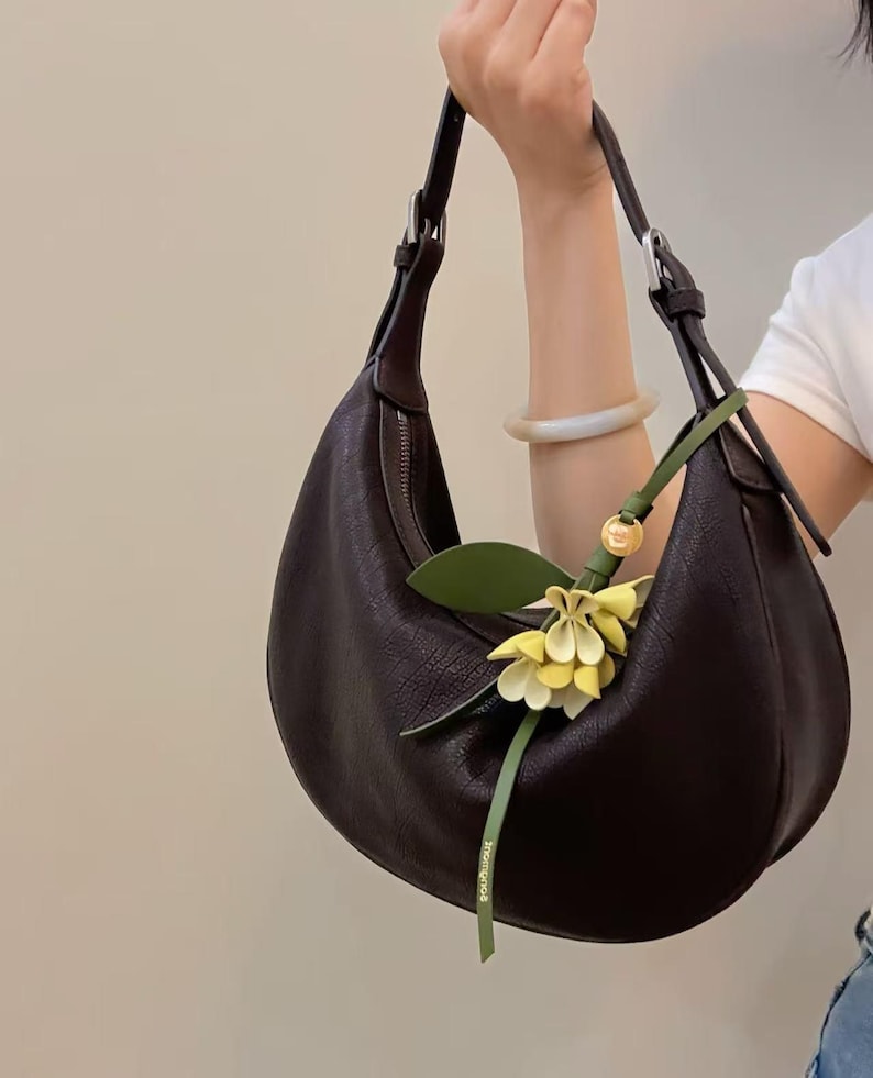 May include: A dark brown crescent-shaped handbag with a textured surface. The bag has a long shoulder strap and a decorative green and yellow floral accent. The bag is held by a person.