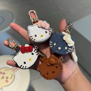 May include: A collection of four Hello Kitty-themed keychains in various colors and materials. The keychains feature the iconic character's face with bows and zipper closures. The keychains are in white, silver, denim blue, and brown, with gold-colored hardware.