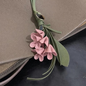 May include: A close-up of a taupe leather bag with a decorative floral tassel. The tassel features pink leather flowers, green leaves, and a green leather strap with gold-tone hardware. The bag has a textured surface.