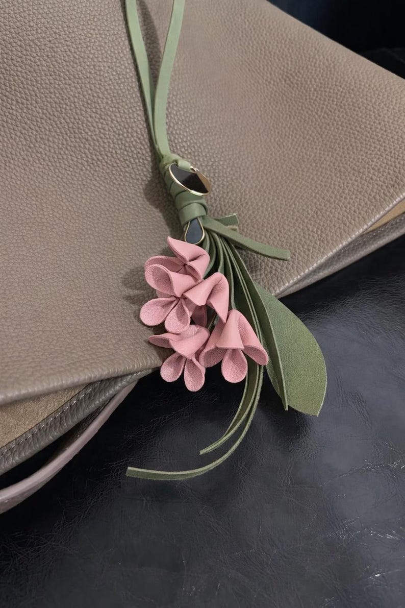 May include: A close-up of a taupe leather bag with a decorative floral tassel. The tassel features pink leather flowers, green leaves, and a green leather strap with gold-tone hardware. The bag has a textured surface.