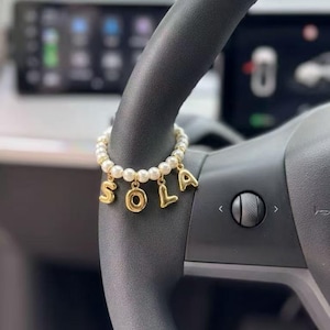 Op de afbeelding: Een parelarmband met gouden letterbedels met de tekst "SOLA" hangt aan een zwart stuurwiel. De armband is gemaakt van kleine, ronde, witte parels en goudkleurige metalen bedels. Het stuurwiel is zwart en heeft een matte afwerking.
