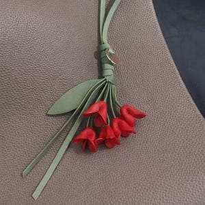 May include: A leather handbag charm featuring a cluster of red tulip-shaped flowers with green stems and leaves. The charm is attached to a light green leather strap and a small gold-colored disc.