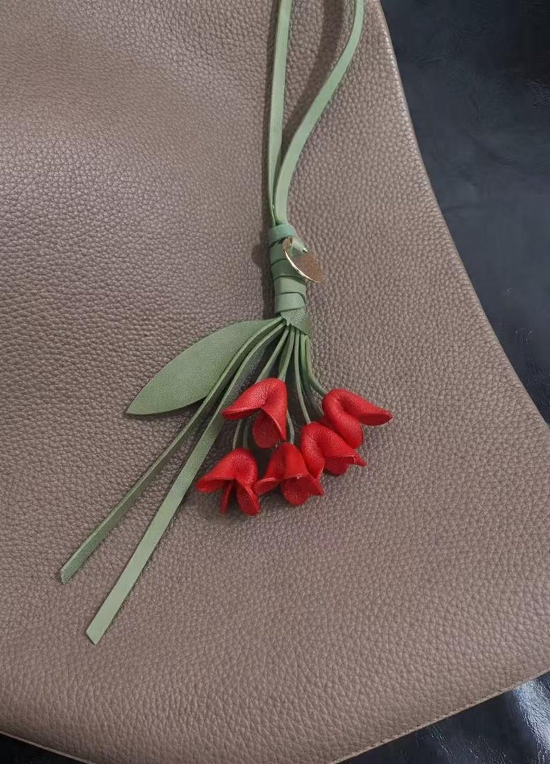 May include: A leather handbag charm featuring a cluster of red tulip-shaped flowers with green stems and leaves. The charm is attached to a light green leather strap and a small gold-colored disc.