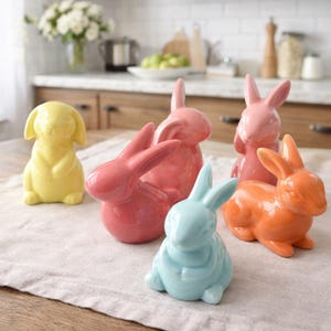 Puede incluir: Colección de figuras de conejos de cerámica coloridas en amarillo, rosa, azul claro y naranja. Los conejos están en varias poses, algunos de pie y otros sentados. Las figuras se exhiben sobre una tela blanca.
