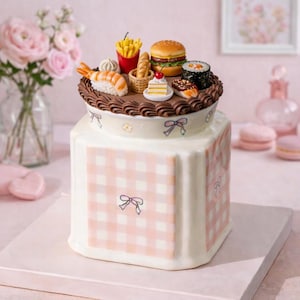 Op de afbeelding: Een decoratieve taart met een witte basis en een roze ruitpatroon. De bovenkant is versierd met miniatuurvoedsel, waaronder een hamburger, sushi, friet en een stuk taart. De taart is versierd met strikken en bloemmotieven.
