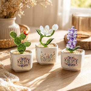 May include: Three glass flower sculptures in small white square pots. The sculptures include a green cactus, a white flower, and a purple flower. Each pot has the word "MOM" in a floral design.