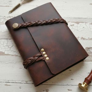 May include: A dark brown leather journal featuring a wrap-around braided leather strap closure secured with a brass button. The cover includes a vertical row of four small brass studs along the edge where the strap fastens, providing a secure hold.