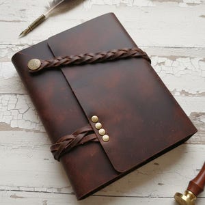 May include: A dark brown leather journal with a braided leather strap closure and brass rivets. A feather quill and a brass stamp are also visible. The journal is closed and appears to be new.