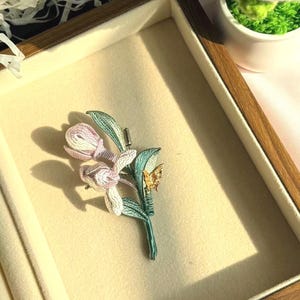 Può includere: Una spilla floreale con due boccioli di fiori avvolti in filo rosa pallido e bianco su uno stelo verde con foglie verdi e un piccolo dettaglio a forma di farfalla dorata. La spilla ha un meccanismo a spillo argentato sul retro dello stelo.