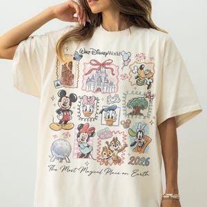 May include: Cream-colored t-shirt featuring a collage of Disney characters and landmarks. The design includes Mickey Mouse, Minnie Mouse, Donald Duck, and Goofy, along with the text "Walt Disney World" and "2026". The shirt also has the phrase "The Most Magical Place on Earth".